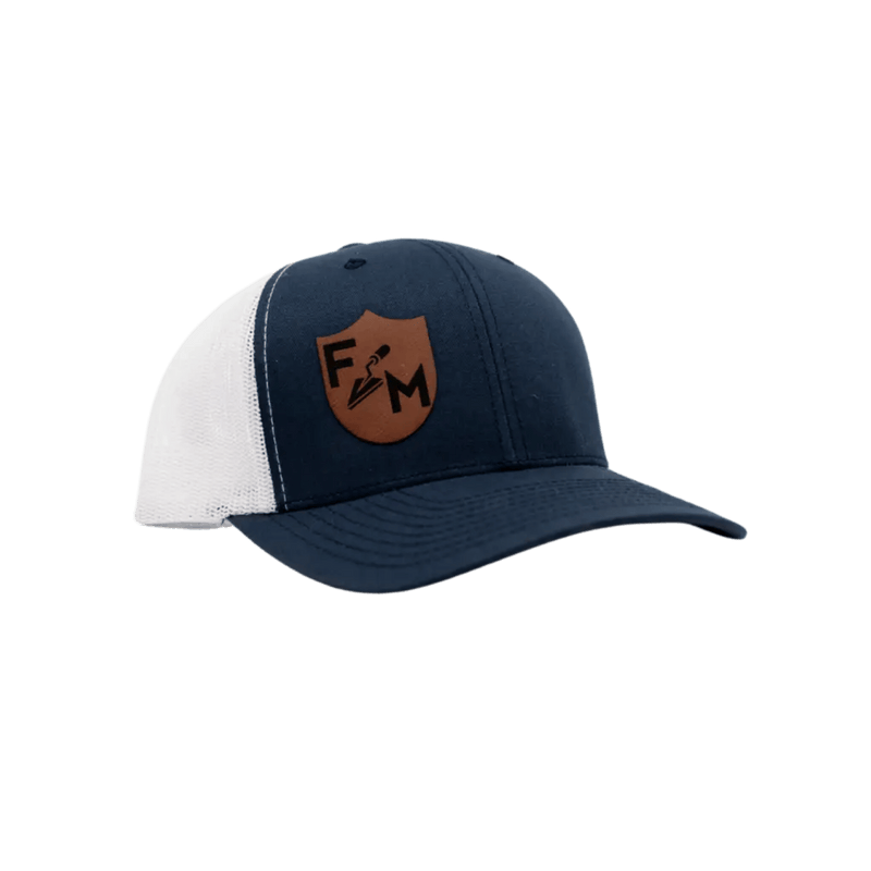 A navy blue and white trucker-style cap with a brown shield patch on the front featuring the letters F and M