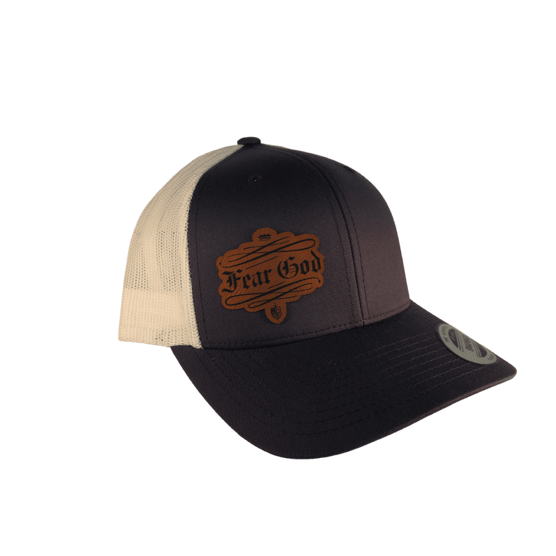 A trucker-style baseball cap with a dark navy front panel and brim, cream mesh back panels, and a brown embroidered 'Fear God' patch on the front, shown against a solid green background.
