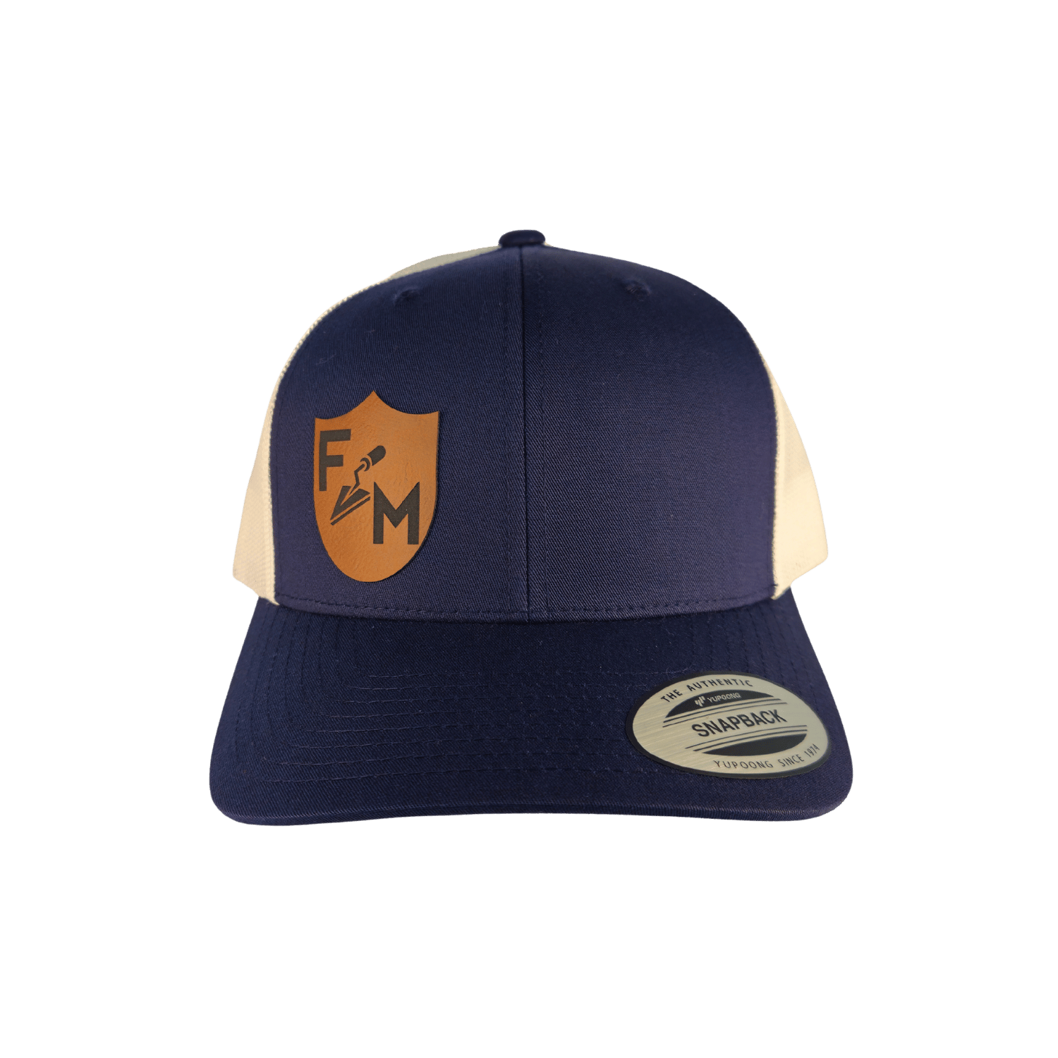 A navy blue and cream trucker hat with a tan shield logo on the front panel. The hat features navy blue front panels and brim, cream mesh back panels, and a silver Snapback sticker on the brim.
