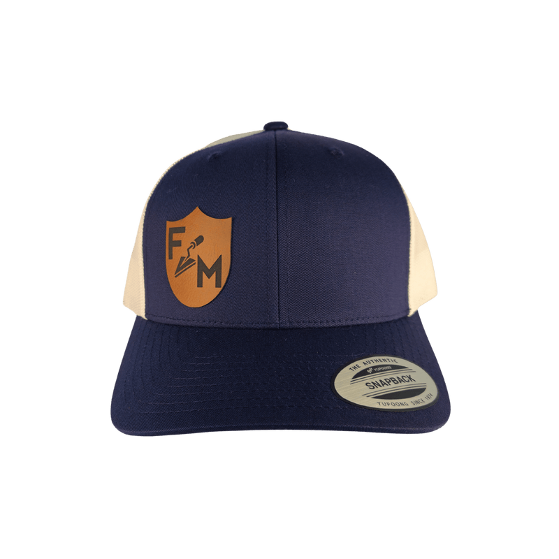 A navy blue and cream trucker hat with a tan shield logo on the front panel. The hat features navy blue front panels and brim, cream mesh back panels, and a silver Snapback sticker on the brim.