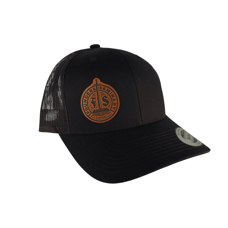 A black trucker-style baseball cap with a curved brim, black mesh back panels, and a circular brown leather patch on the front that reads 'Founders Seminary' with an 'FS' emblem. A silver-toned metal sticker is visible on the brim.