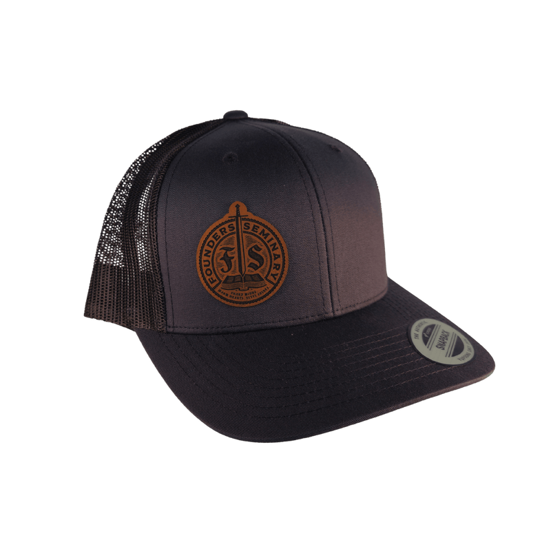 A dark charcoal trucker hat with black mesh back panels and a brown leather patch on the front.