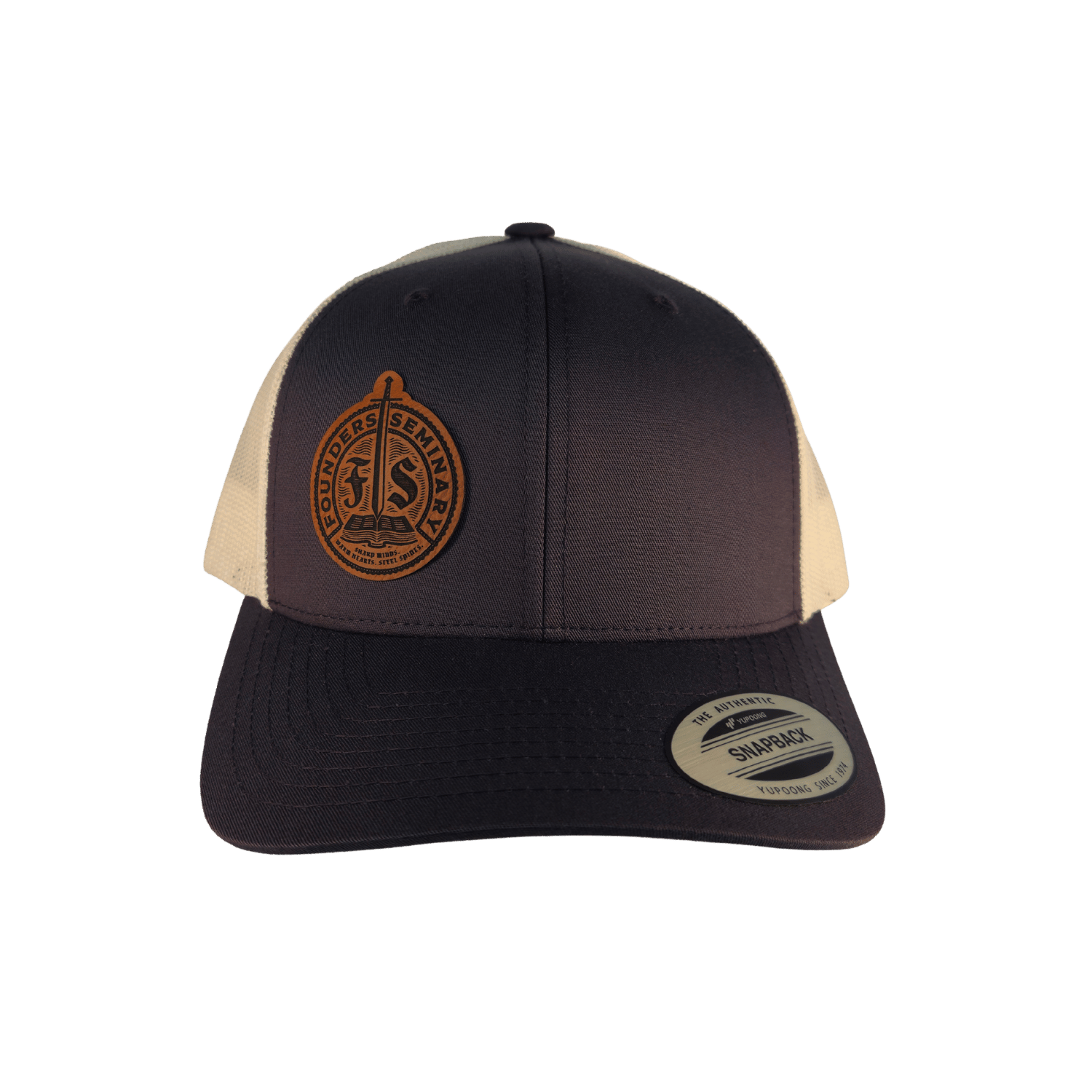A structured trucker-style baseball cap with a dark grey front and brim, cream mesh side panels, and a circular brown leather patch on the front. 