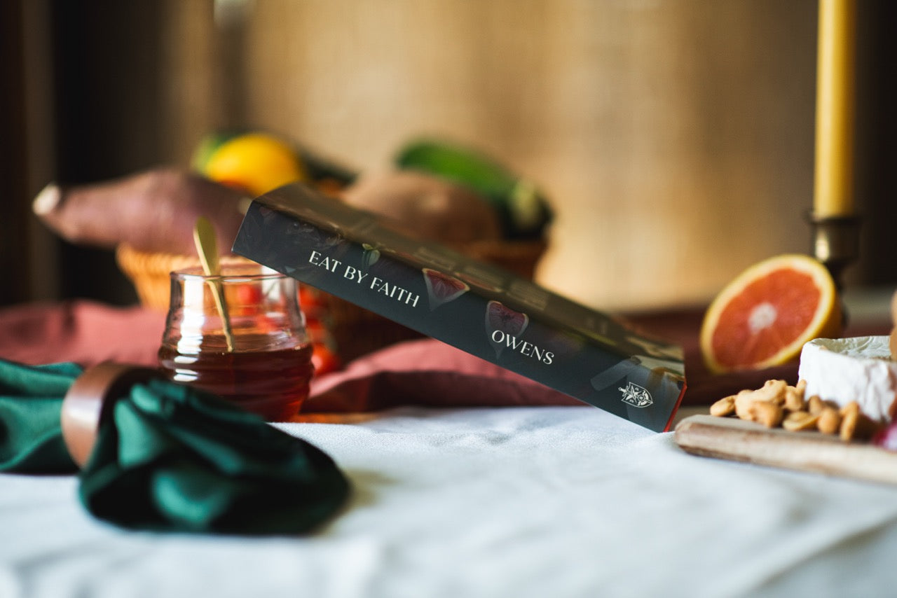 A book titled 'Eat by Faith' by Owens lies diagonally on a white tablecloth, surrounded by a styled rustic spread with a glass jar of honey, a green napkin, nuts, cheese, and fruit in a warm, softly blurred background.