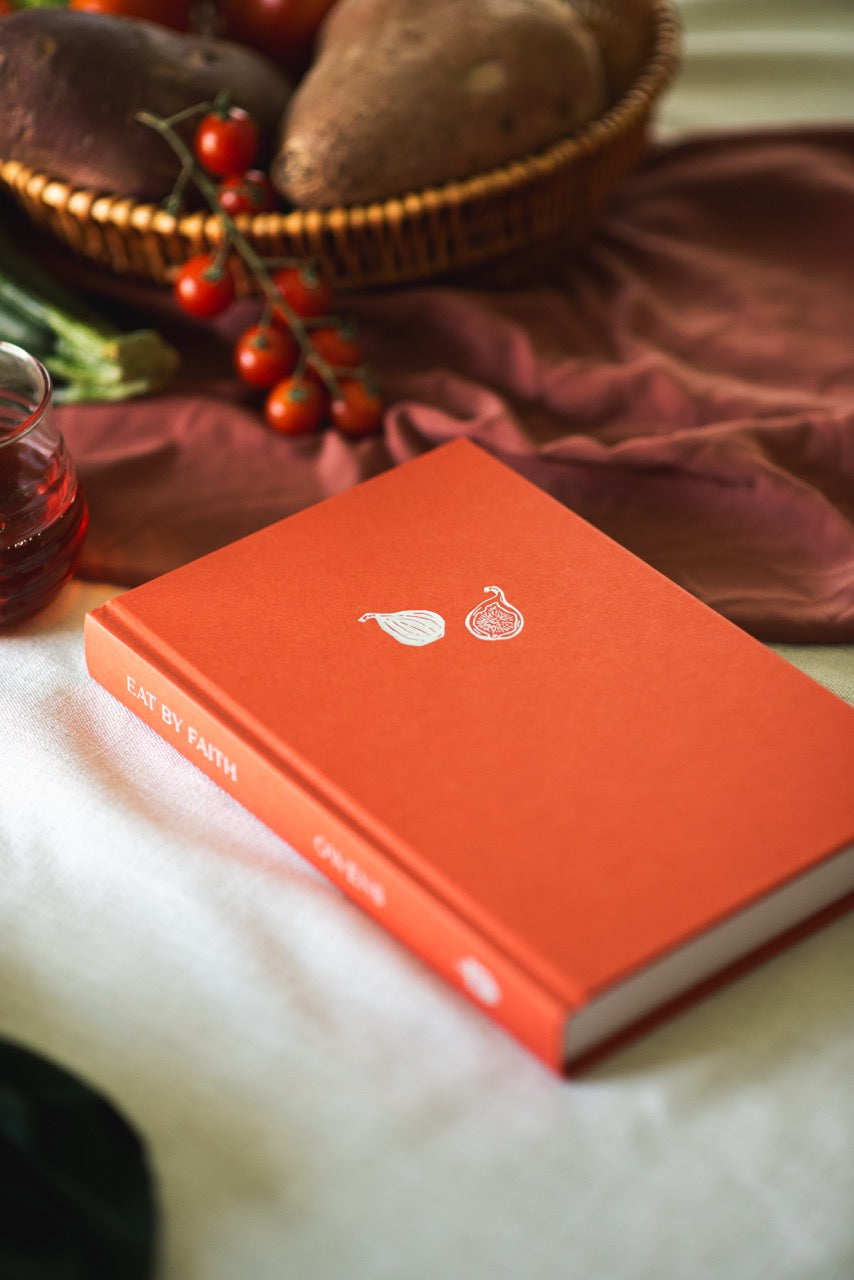 An orange hardcover book titled 'Eat of Earth' lies on a white fabric surface, styled with a basket of sweet potatoes, red berries, and draped burgundy cloth in the background.