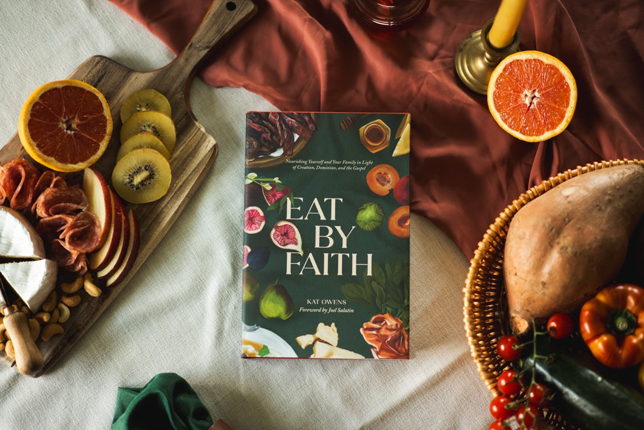A book titled "Eat By Faith" by Kat Owens is displayed in a styled flat-lay setting with food props including sliced oranges, kiwi, cheese, crackers, a sweet potato, and vegetables on a draped fabric background.