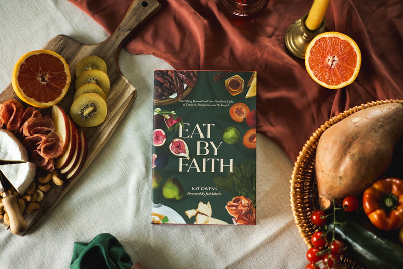 A book titled "Eat By Faith" by Kat Owens is displayed in a styled flat-lay setting with food props including sliced oranges, kiwi, cheese, crackers, a sweet potato, and vegetables on a draped fabric background.