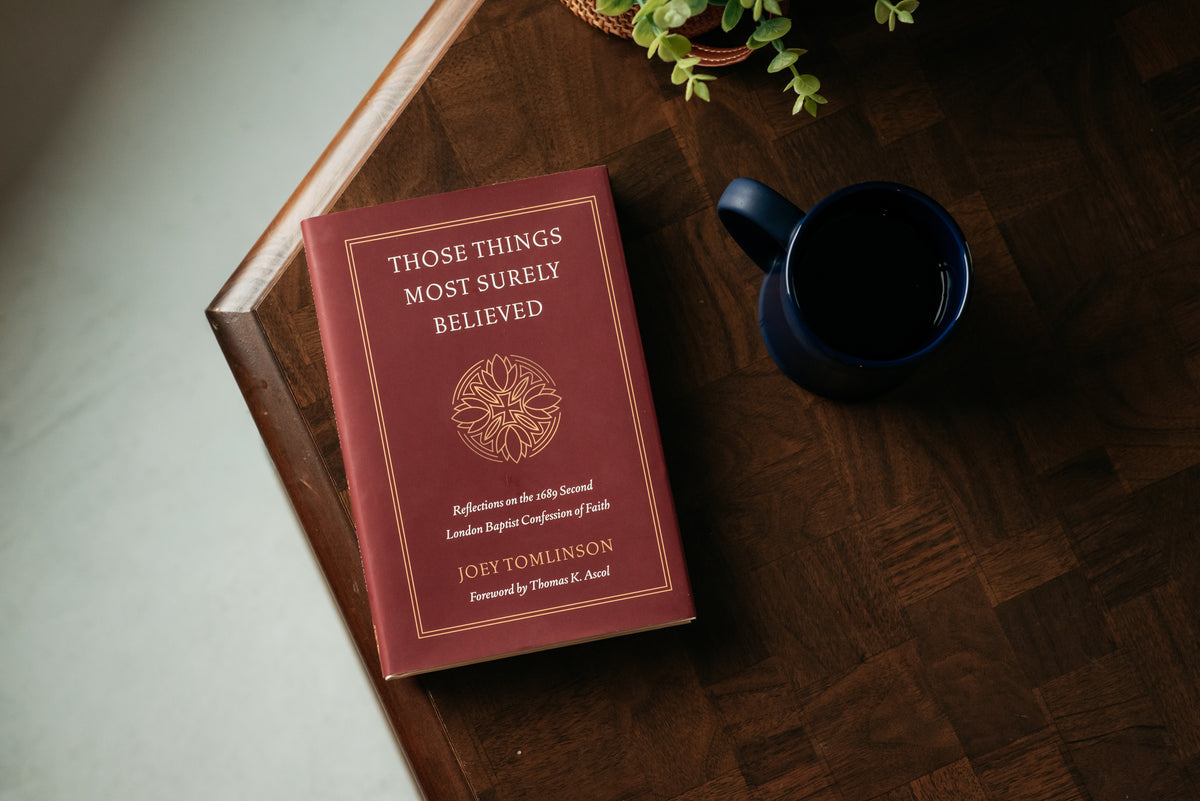 A hardcover book titled 'Those Things Most Surely Believed' by Joey Tomlinson, placed on top of white books on a dark wood surface. The book cover is deep burgundy with gold text and a circular emblem.