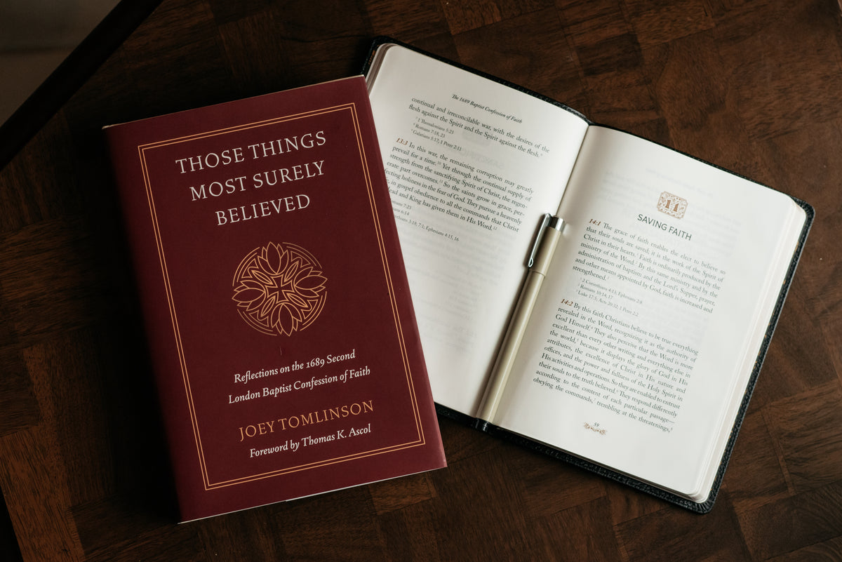 A hardcover book titled 'Those Things Most Surely Believed' by Joey Tomlinson, placed on top of white books on a dark wood surface. The other book is an opened copy of the leather 1689 to Chapter 14