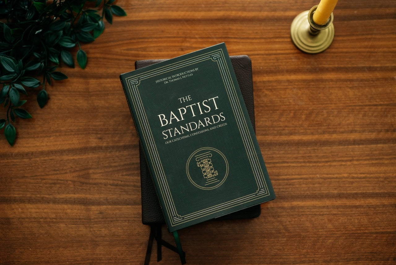 A dark green hardcover book titled 'The Baptist Standards' placed on a wooden tabletop, with a candle holder and green leaves partially visible in the scene.
