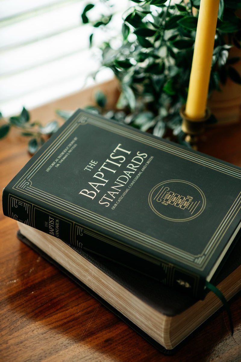 A stack of two hardcover books on a wooden table, with the top book titled "The Baptist Standards" in dark green with gold lettering. A yellow candlestick and green foliage are softly blurred in the background.