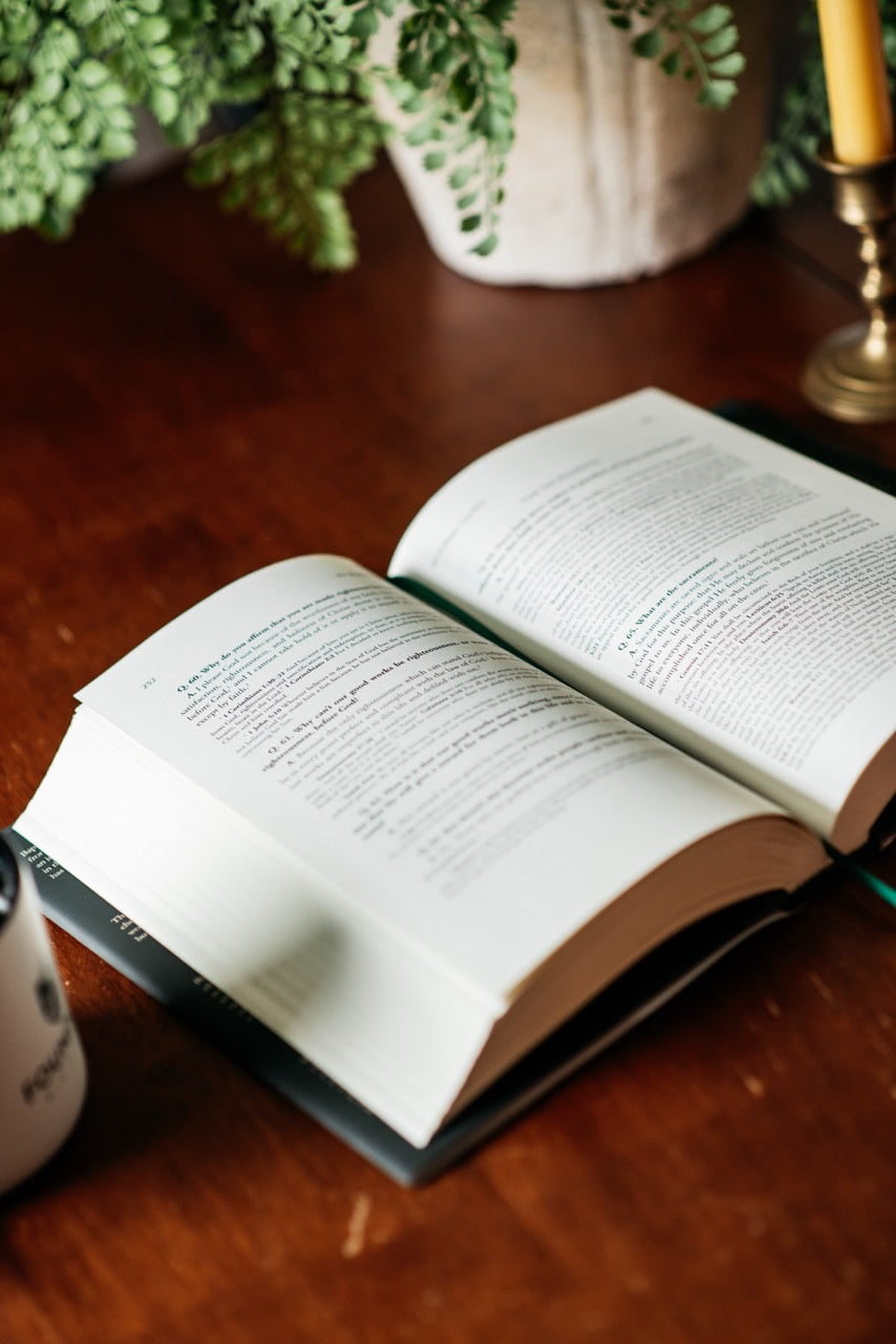 An open book with printed pages lies on a wooden table, surrounded by soft-focus props including greenery, a candle, and part of a mug.
