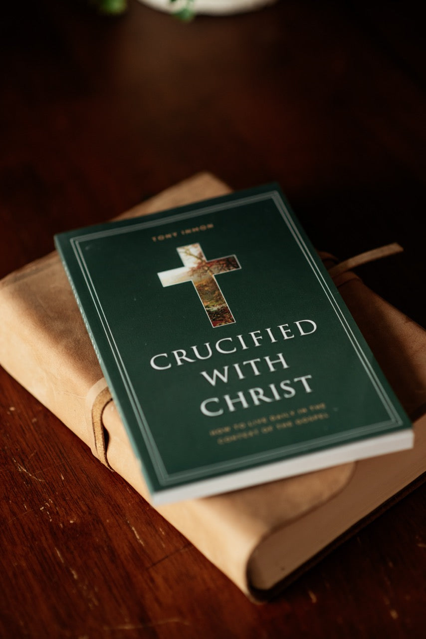 A green book titled 'Crucified with Christ' by Tony Inmon, featuring a cross emblem on the cover, resting on a tan leather-bound journal over a dark wooden surface.