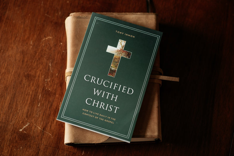 A book titled "Crucified with Christ" by Tony Inmon lies on top of a brown cloth-covered book on a dark wooden table. The book cover is dark green with a decorative cross and white text.