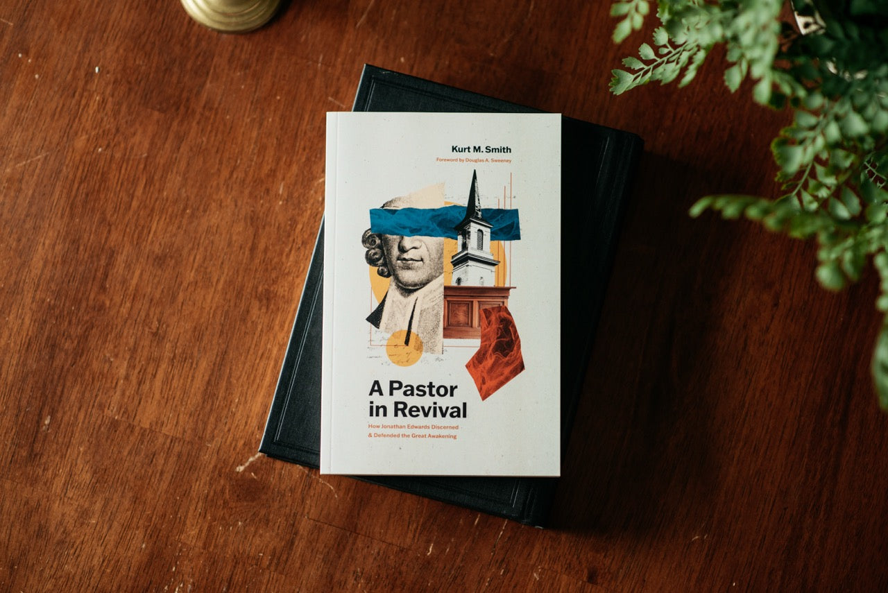 A book titled "A Pastor in Revival" by Kurt M. Smith lies on top of a black book on a wooden tabletop, with a green plant leaf visible at the right edge.