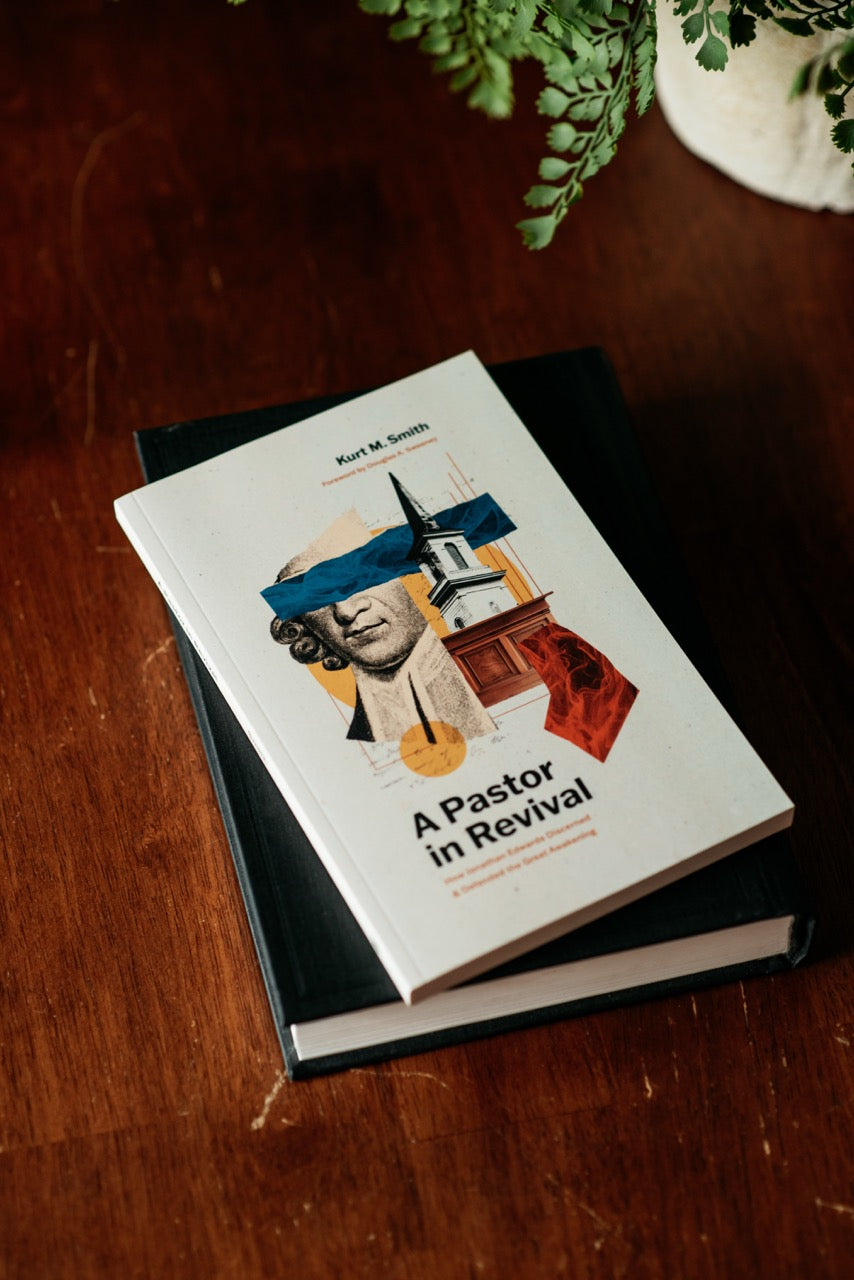 A hardcover book titled "A Pastor in Revival" by Kurt M. Smith lies on top of a black book on a dark wooden table.