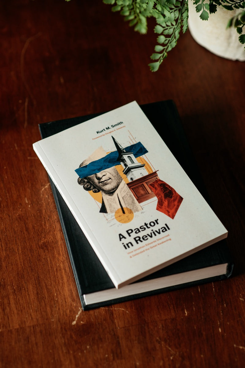 A hardcover book titled "A Pastor in Revival" by Kurt M. Smith lies on top of a black book on a dark wooden table.