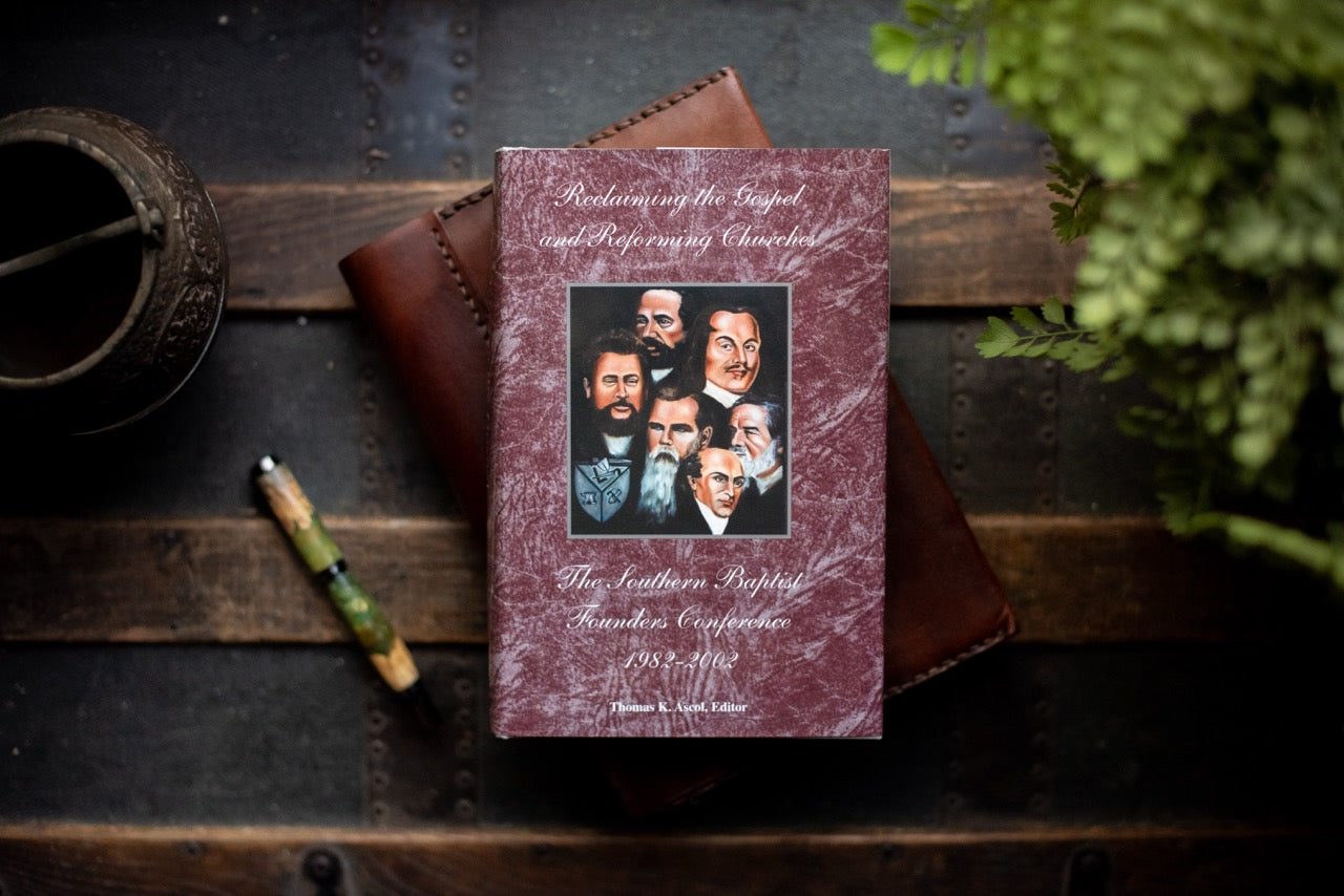 Book titled 'Reclaiming the Gospel and Reforming the Church lies on a dark wooden surface with a brown leather wallet, a small metal container, a pen, and green foliage around it.
