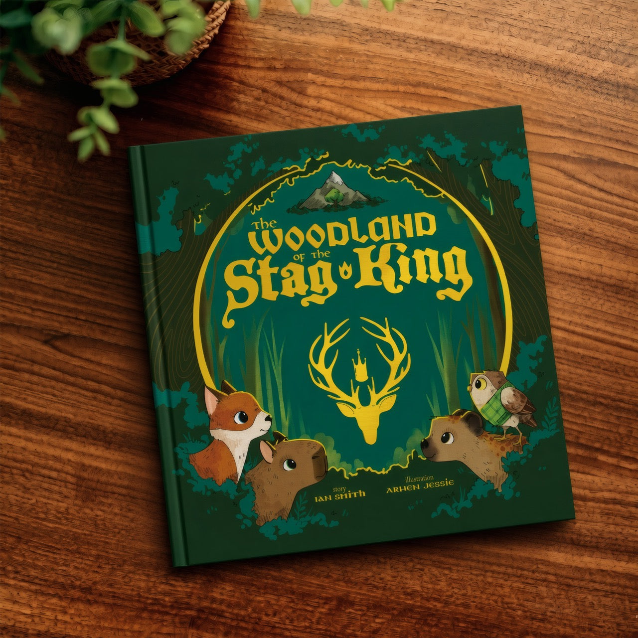 A hardcover children's book titled "The Woodland of the Stag King" lies on a wooden tabletop. The cover is dark green with yellow title lettering, a stag silhouette, and illustrated woodland animals including a fox, rabbit, owl, and bird.