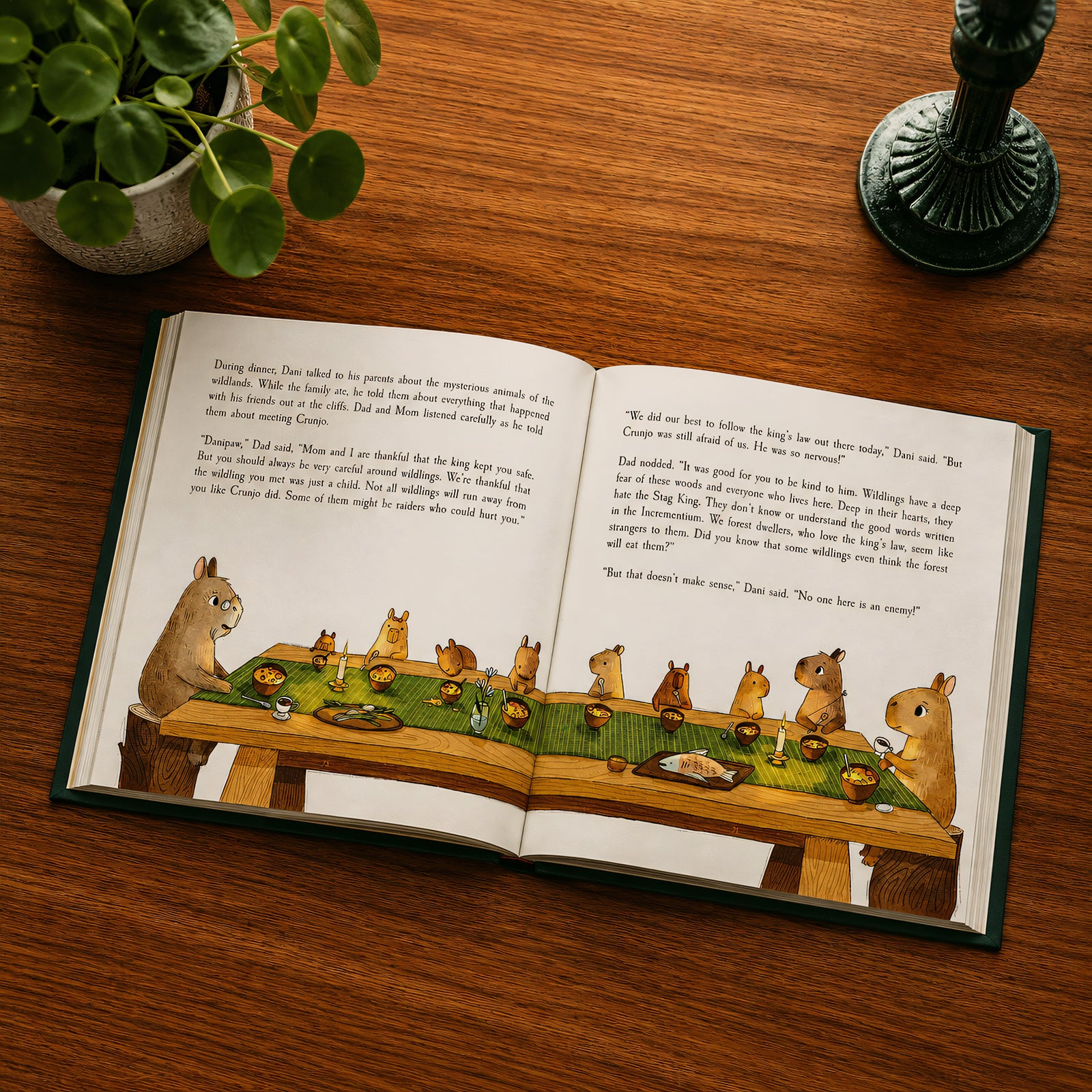 An open children's book with illustrated woodland animals sitting around a table, photographed from above on a wooden surface with a potted plant and candlestick nearby.
