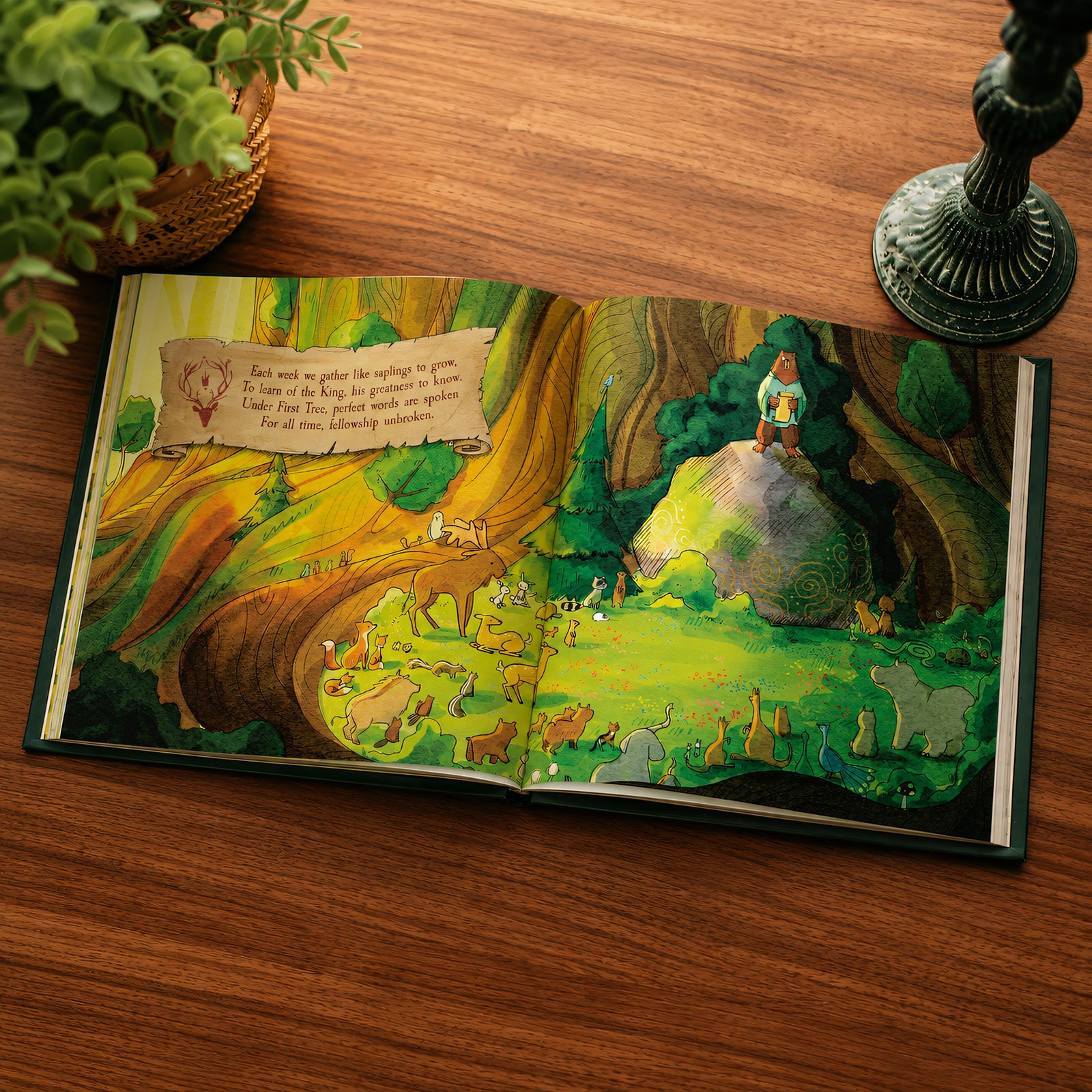 An open illustrated children's book spread on a wooden table, showing a colorful forest scene with animals and a small character standing on a rock. A potted plant and a dark decorative object are partially visible around it.