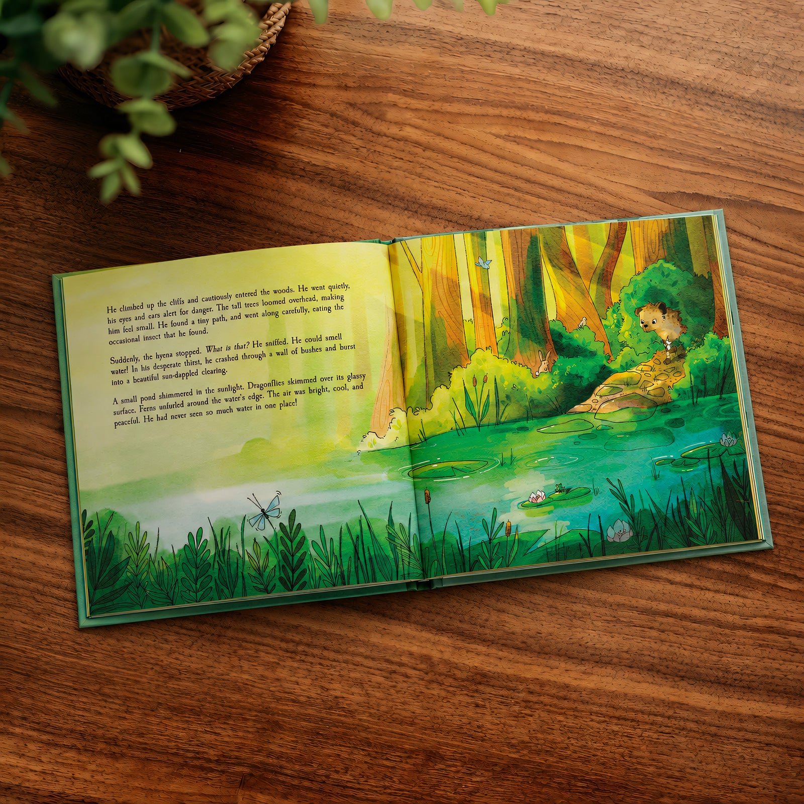 An open children's picture book lying on a wooden tabletop, showing a colorful forest scene with a pond, trees, and a girl by the water. The left page has printed text, and the right page shows an illustrated woodland landscape.