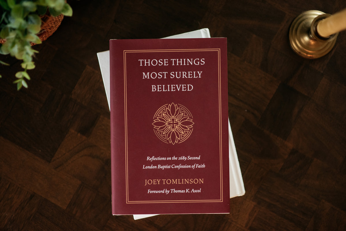 A hardcover book titled 'Those Things Most Surely Believed' by Joey Tomlinson, placed on top of white books on a dark wood surface. The book cover is deep burgundy with gold text and a circular emblem.
