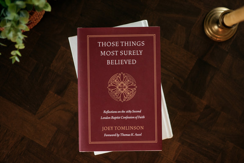 A hardcover book titled 'Those Things Most Surely Believed' by Joey Tomlinson, placed on top of white books on a dark wood surface. The book cover is deep burgundy with gold text and a circular emblem.