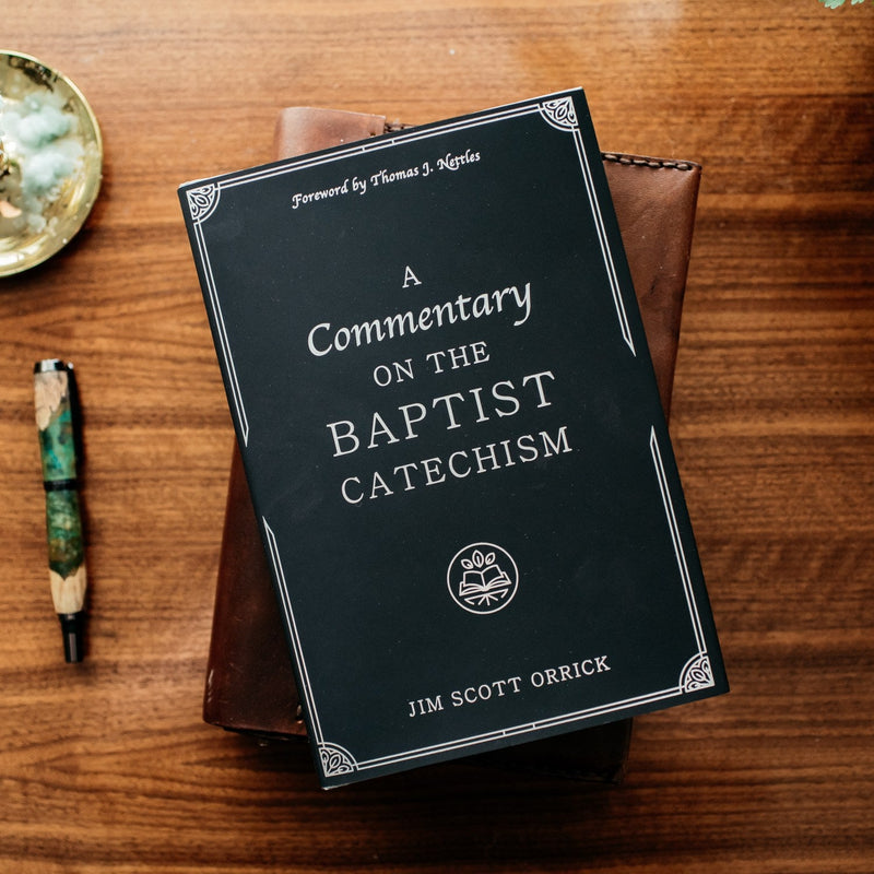 Book titled 'A Commentary on the Baptist Catechism' by Jim Scott Orrick on a wooden surface with a pen and decorative item.