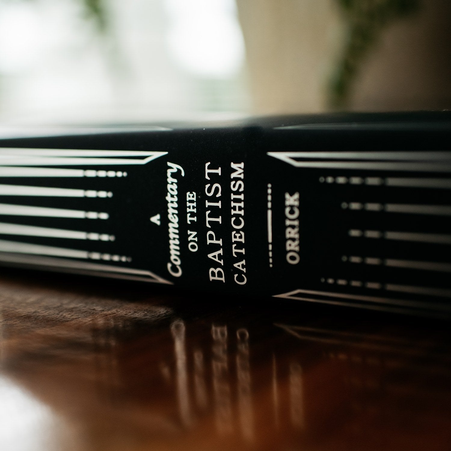 Book titled 'A Commentary on the Baptist Catechism' by Orrick on a wooden surface with a blurred background.