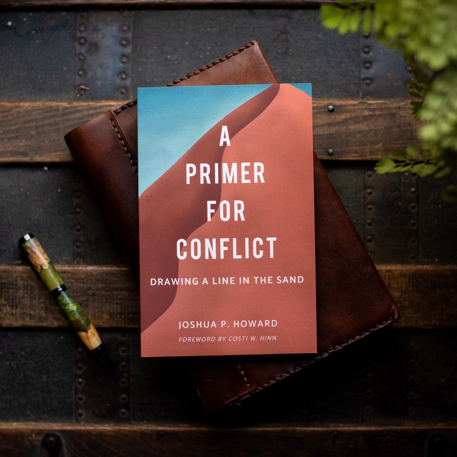 Book titled 'A Primer for Conflict' by Joshua P. Howard on a wooden surface with a pen and plant.