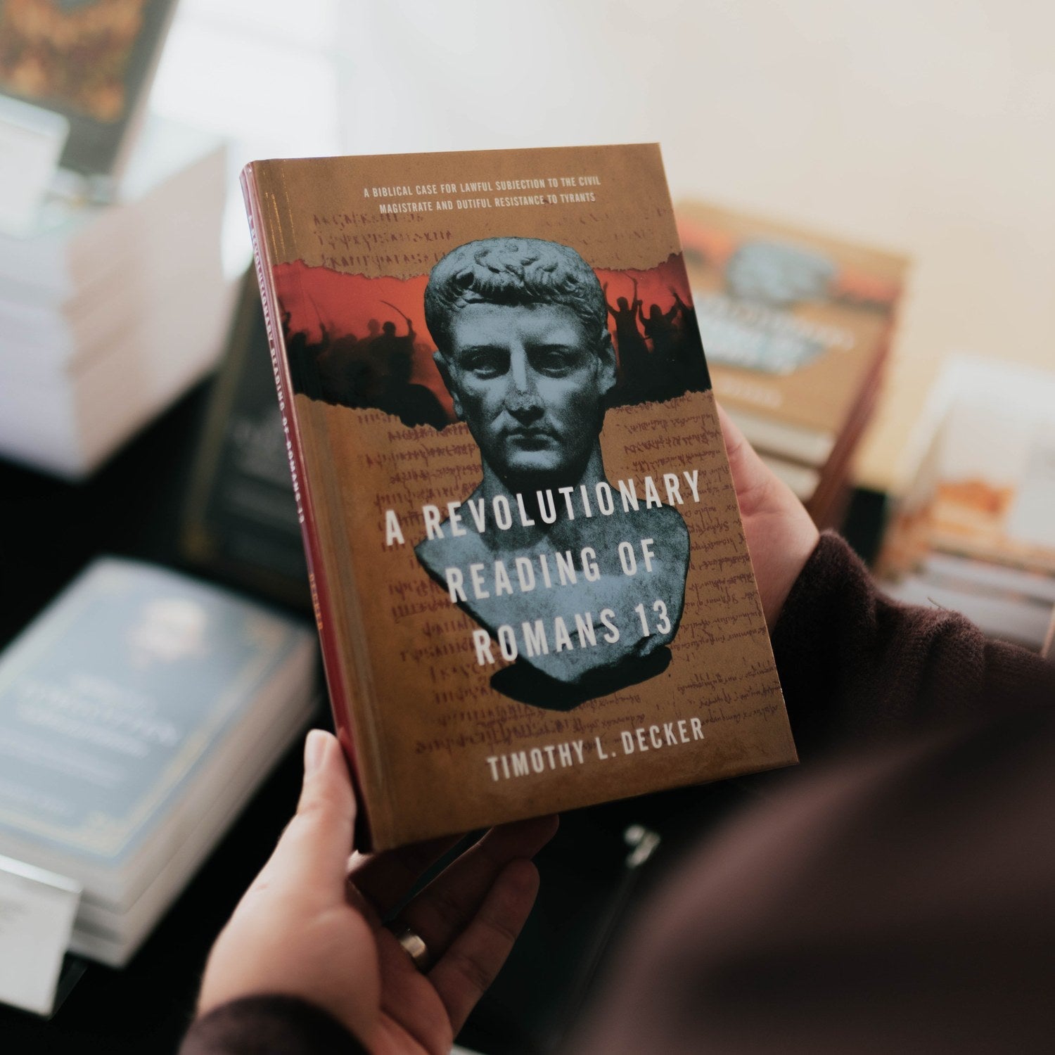 Book titled 'A Revolutionary Reading of Romans 13' held by a person with blurred bookshelf in the background