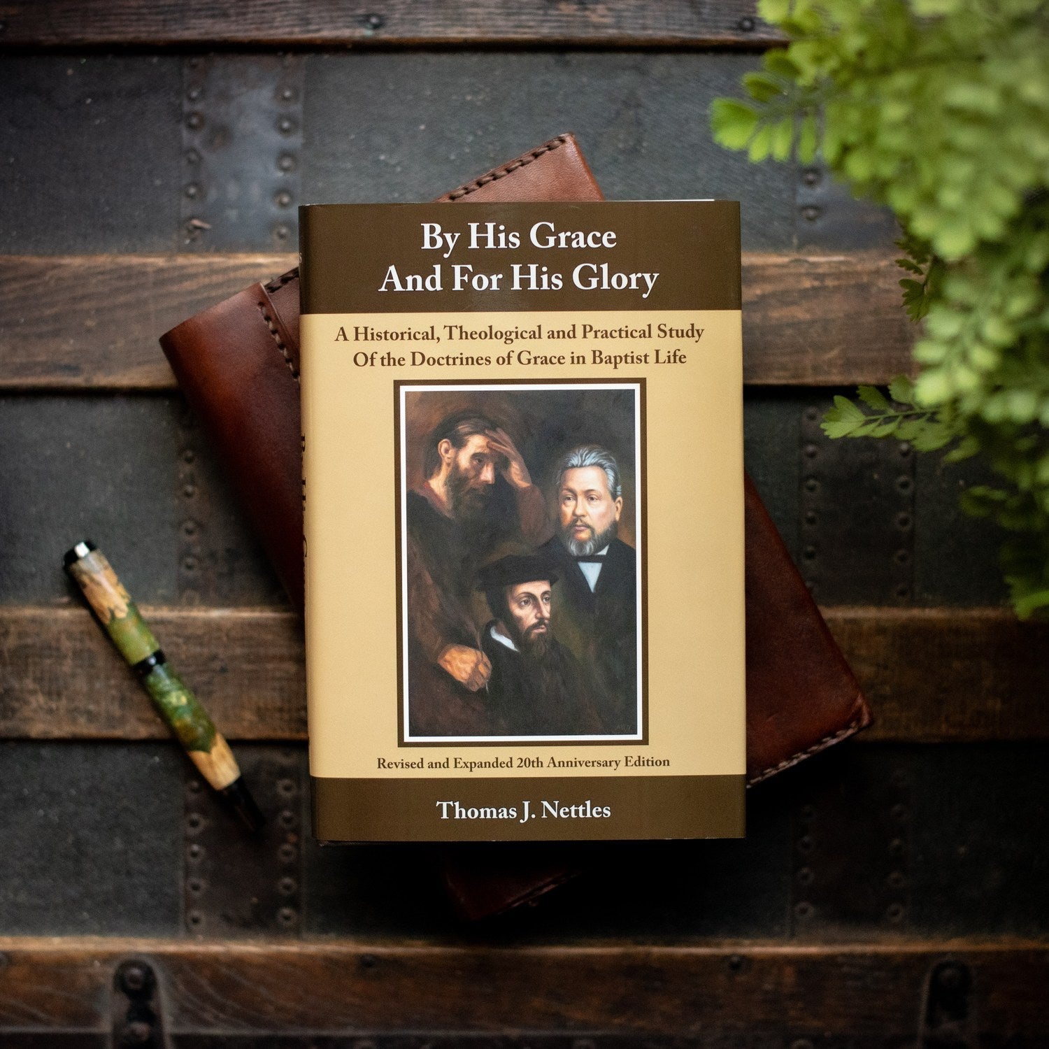 Book titled 'By His Grace And For His Glory' by Thomas J. Nettles on a wooden surface with a pen and plant.