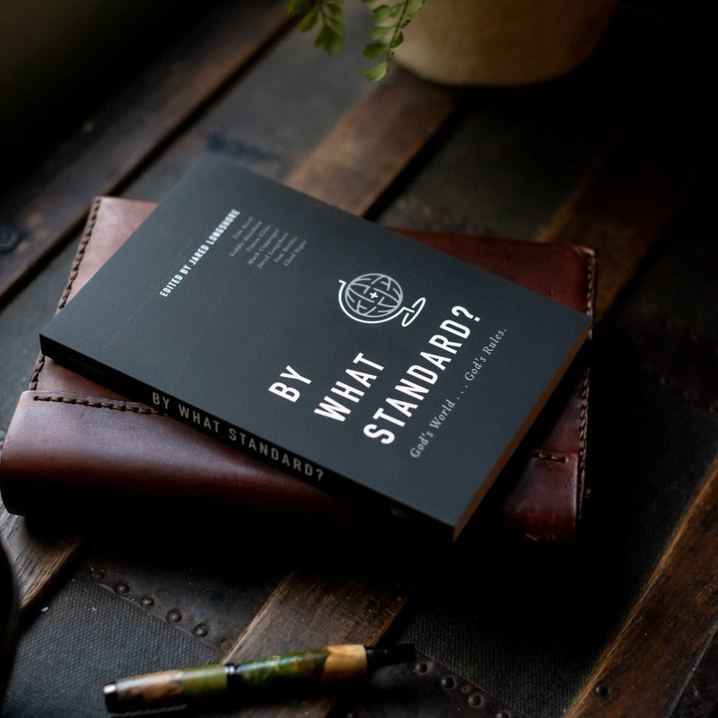 Book titled 'By What Standard?' on a wooden surface with a pen and plant in the background