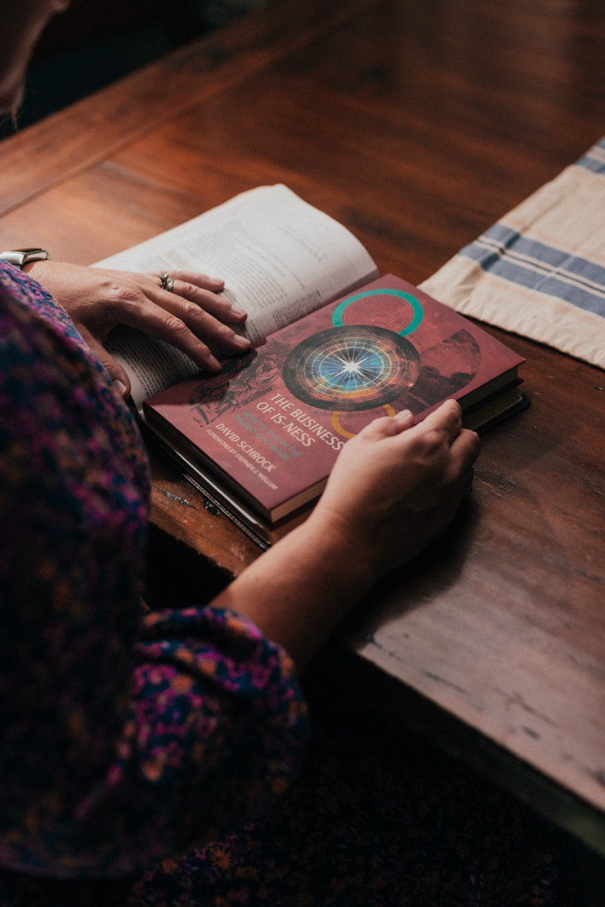 A person's hands resting on a stack of books on a rustic wooden table, with the top book titled 'The Business of Is-Ness' visible.