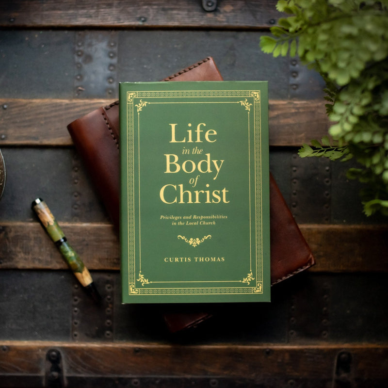 Book titled 'Life in the Body of Christ' by Curtis Thomas on a wooden surface with a pen and leather item.