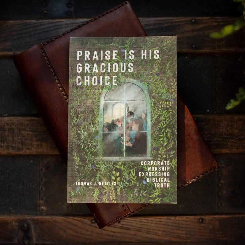 Book titled 'Praise is His Gracious Choice' by Thomas J. Nettles on a wooden surface with a brown leather object.