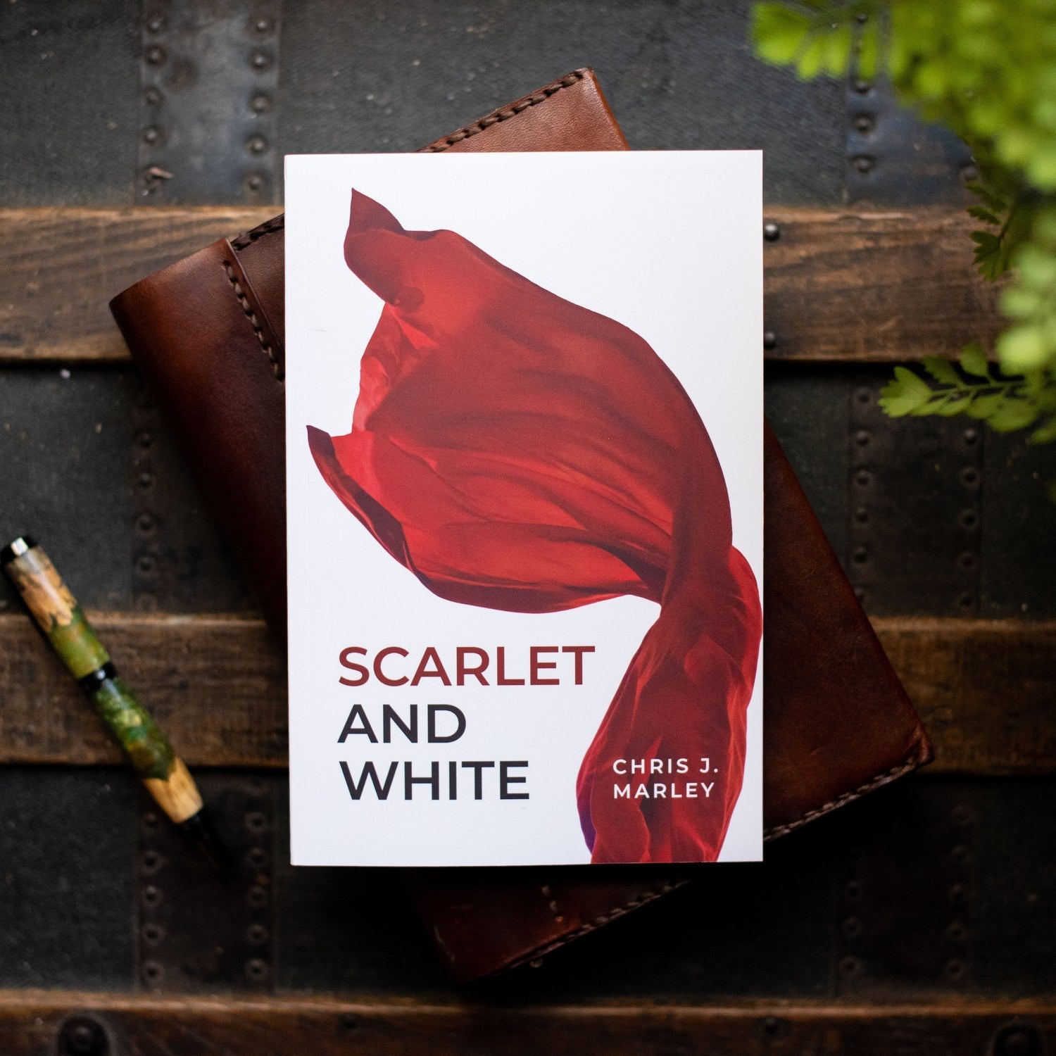 Book cover of 'Scarlet and White' by Chris J. Marley on a wooden surface with a pen and leather journal.