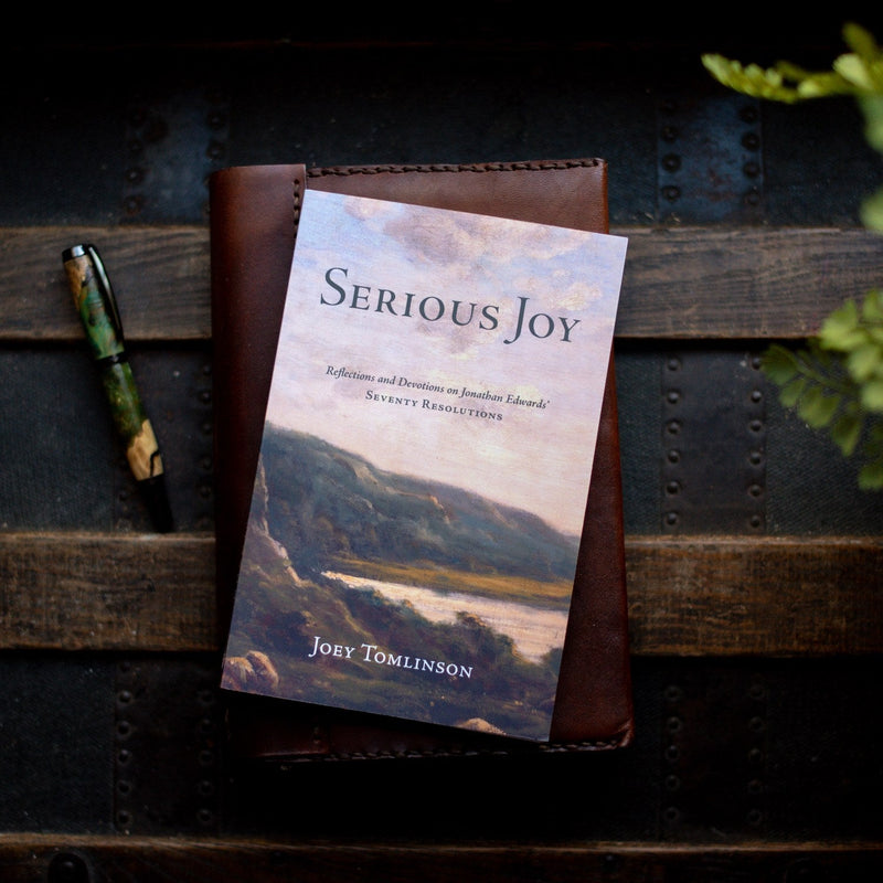 Book titled 'Serious Joy' by Joey Tomlinson on a wooden surface with a pen and plant.