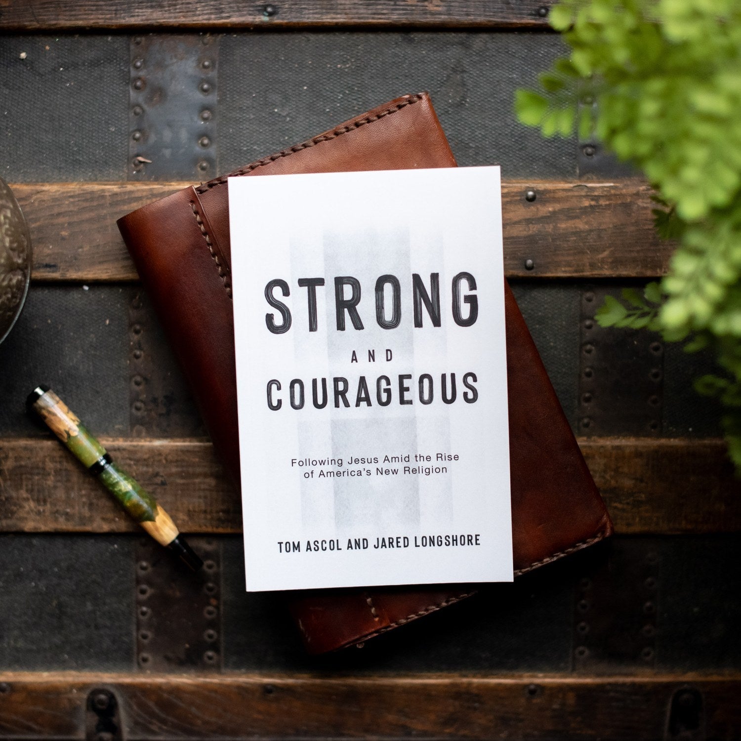 Book titled 'Strong and Courageous' by Tom Ascol and Jared Longshore on a wooden surface with a pen and plant.