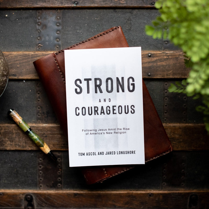 Book titled 'Strong and Courageous' by Tom Ascol and Jared Longshore on a wooden surface with a pen and plant.