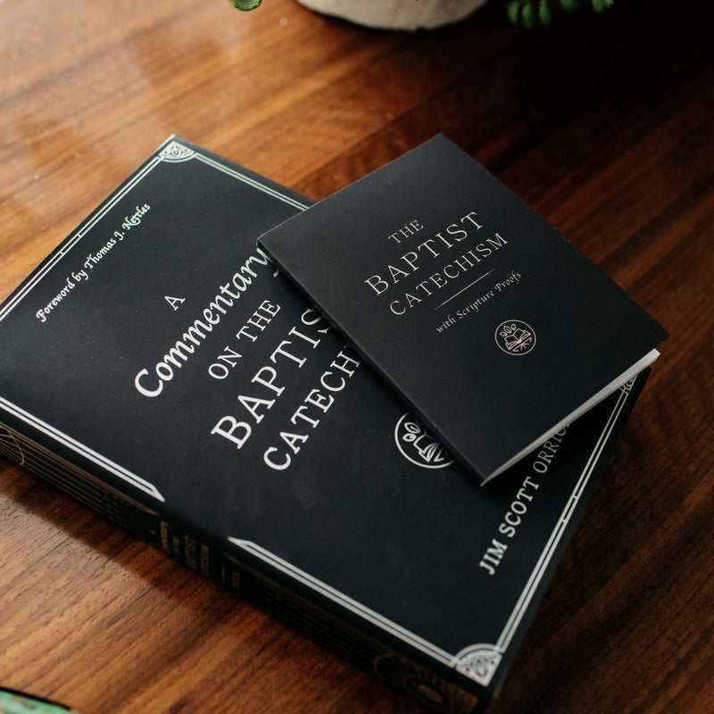 Two black books titled 'A Commentary on the Baptist Catechism' and 'The Baptist Catechism' on a wooden surface.